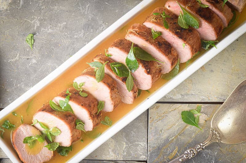 Sliced pork tenderloin with a wine and orange sauce in a white rectangular dish, large spoon on the side, tile background.