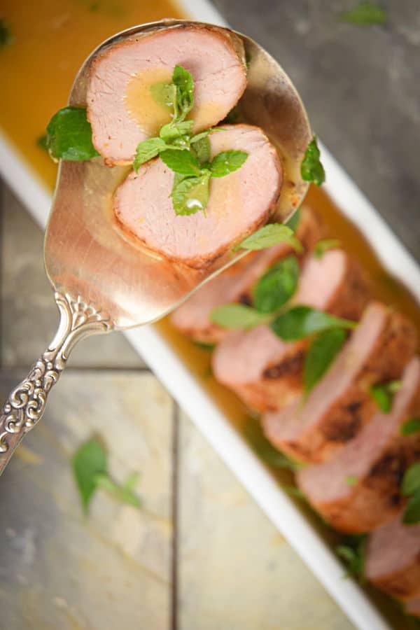 Sliced pork tenderloin with a wine and orange sauce in a white rectangular dish, large spoon on the side, tile background.