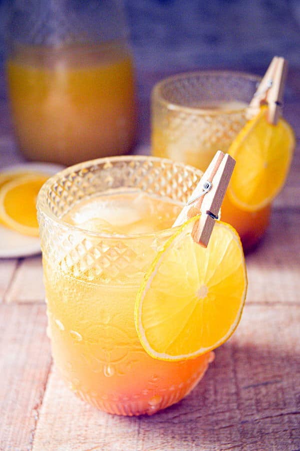 Fall Pumpkin Party Punch in a clear glass with a thin orange slice garnish clipped on with a clothespin, glass pitcher of punch in the background.