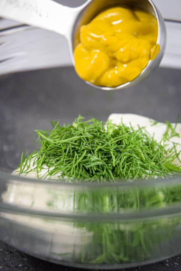 A measuring spoon with mustard in it, over a bowl of mayo and dill.