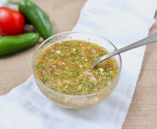 Jalapeno salsa in a clear glass jar with a spoon in it, on top of a white cloth, wooden background.