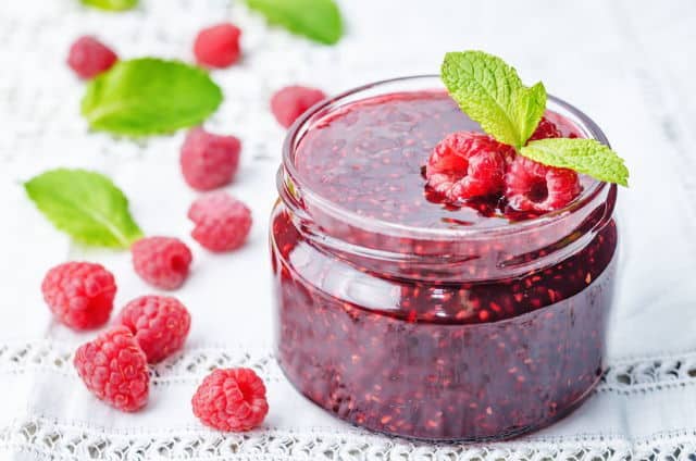 Raspberry jam on a white background, with raspberries on the side.