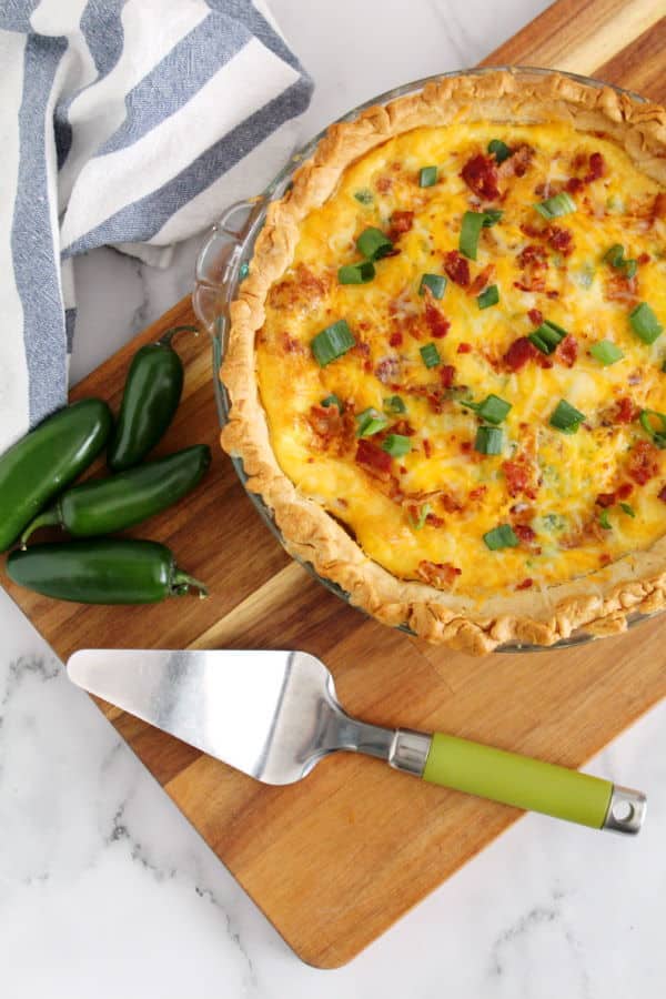 Jalapeno popper quiche in a pie pan on wooden cutting board, pie utensil and jalapenos on the side.