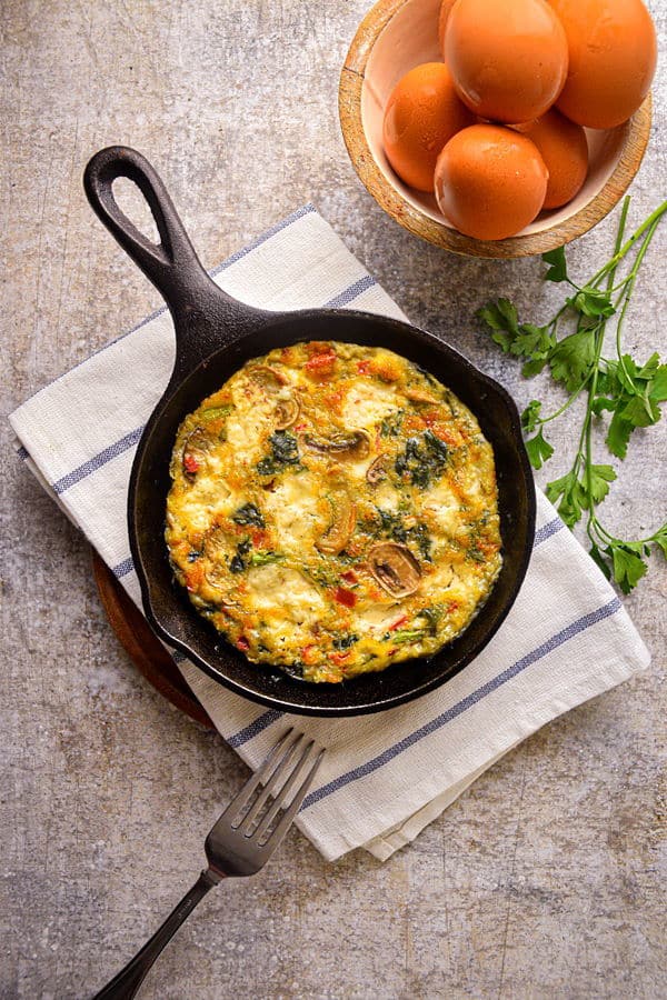 Electric Skillet Vegetable and Goat Cheese Frittata in a cast iron pan, with eggs and cilantro on the side.