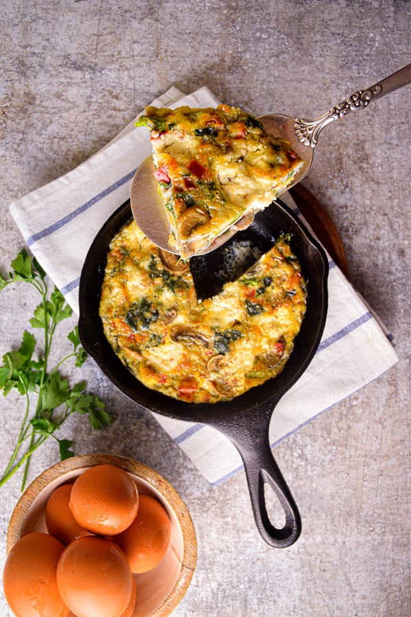 A piece of Electric Skillet Vegetable and Goat Cheese Frittata on a pie server and in a cast iron pan, with eggs and cilantro on the side.