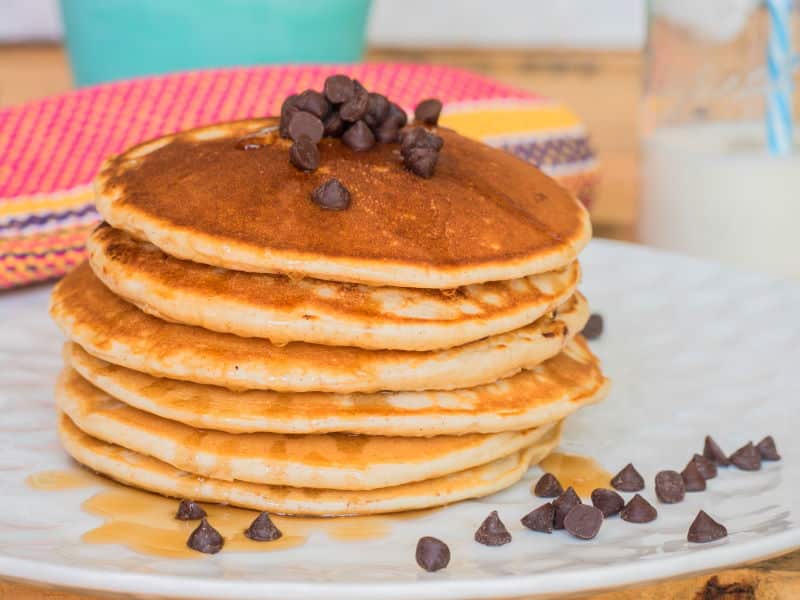 Electric Skillet Pancakes 3 - Inthekitch Pancakes piled on a plate with chocolate chips and maple syrup.