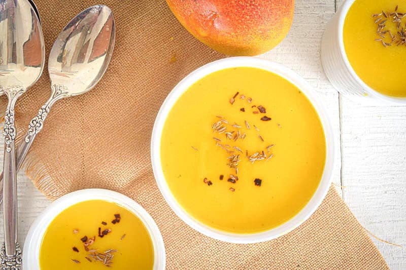 Mango soup in white bowls, 2 spoons and a fresh mango on a brown piece of cloth, white wooden background.