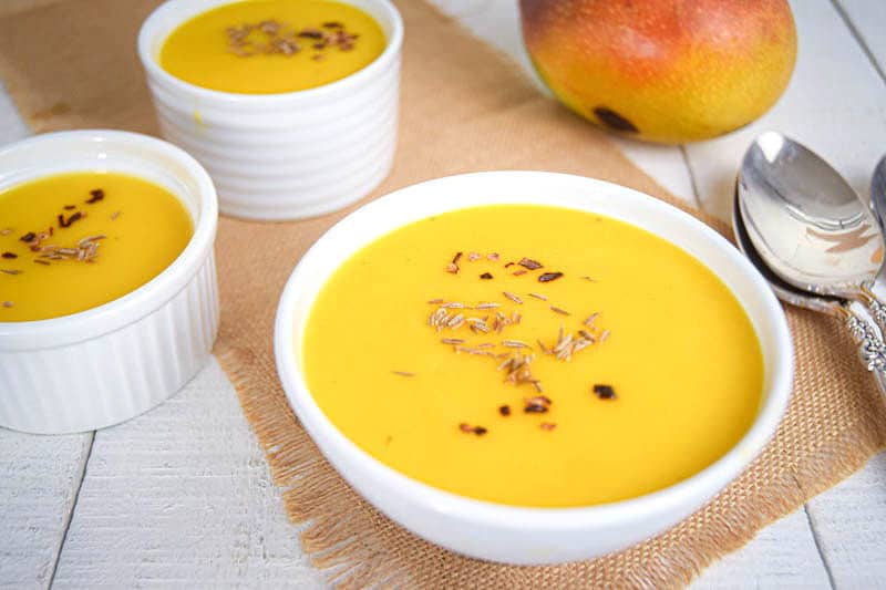 Mango soup in white bowls, 2 spoons and a fresh mango on a brown piece of cloth, white wooden background.