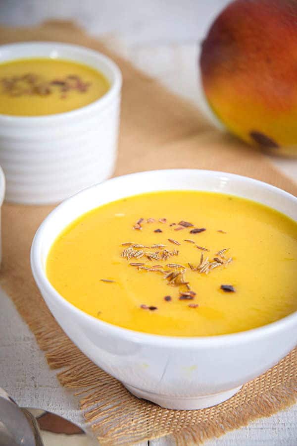 Mango soup in white bowls and a fresh mango on a brown piece of cloth, white wooden background.