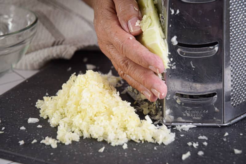 Shredding white cabbage with box grater on dark cutting board.