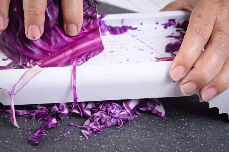How to Shred Cabbage_26 Shredding red cabbage with mandolin on dark cutting board.