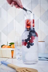 A glass pitcher filled with ice and berries. A measuring spoon of sugar held by a hand.