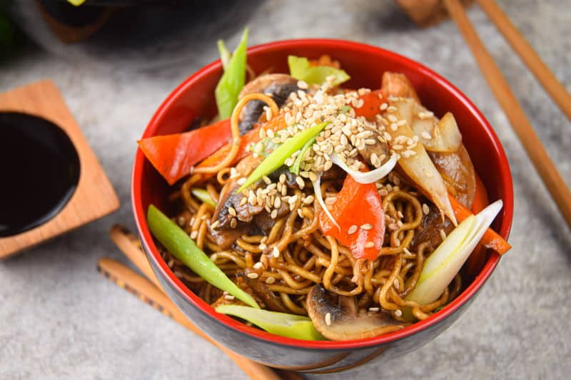 Chicken and vegetable lo mein in red and black bowl, chopsticks and green onions on the side.