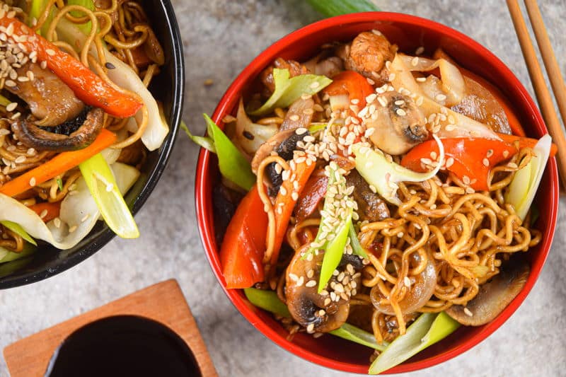 Chicken and vegetable lo mein in red and black bowls, chopsticks and green onions on the side.