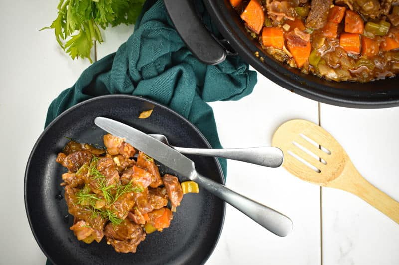 St. Pattys Beef Stew with Black Beer on a black dish, a wooden spoon on the side, white wooden background.