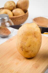 A potato on a wooden cutting board, potatoes in the background.