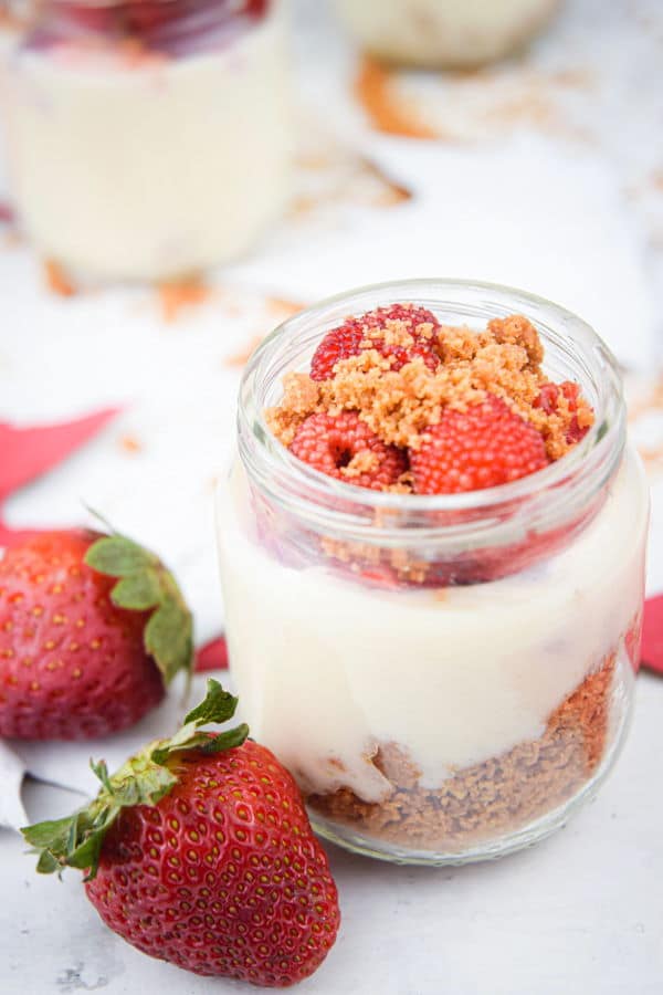 Raspberry cheesecake in mini dessert jars, strawberries in the background.