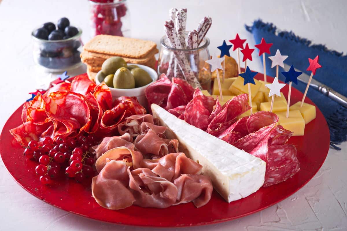 Charcuterie board with red, white and blue colored meats, cheeses, crackers and fruit on red plate.