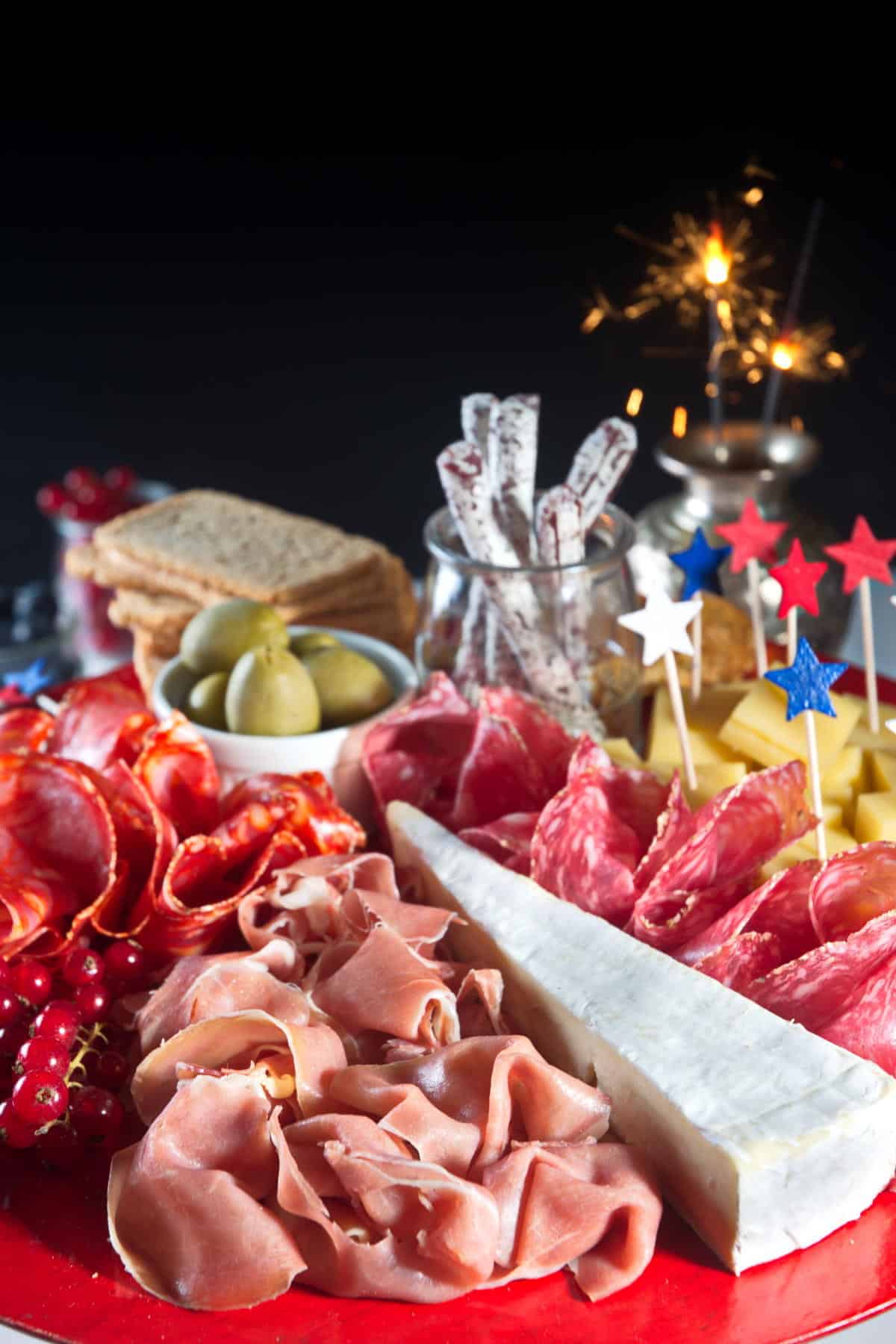 Charcuterie board with red, white and blue star toothpicks on red plate, sparklers lit in the background.