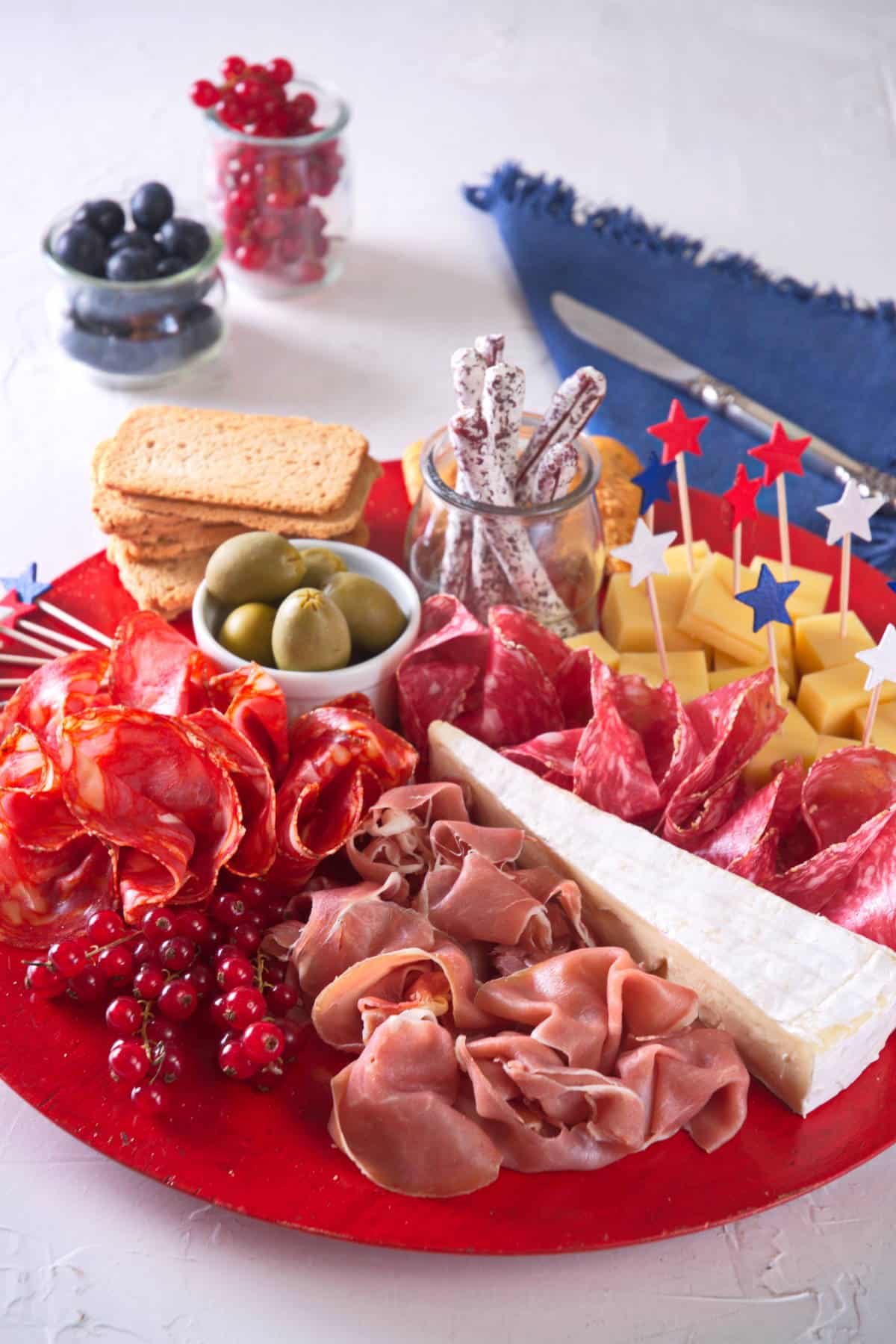 Charcuterie board with red, white and blue colored ingredients on red plate, blue napkin on the side.