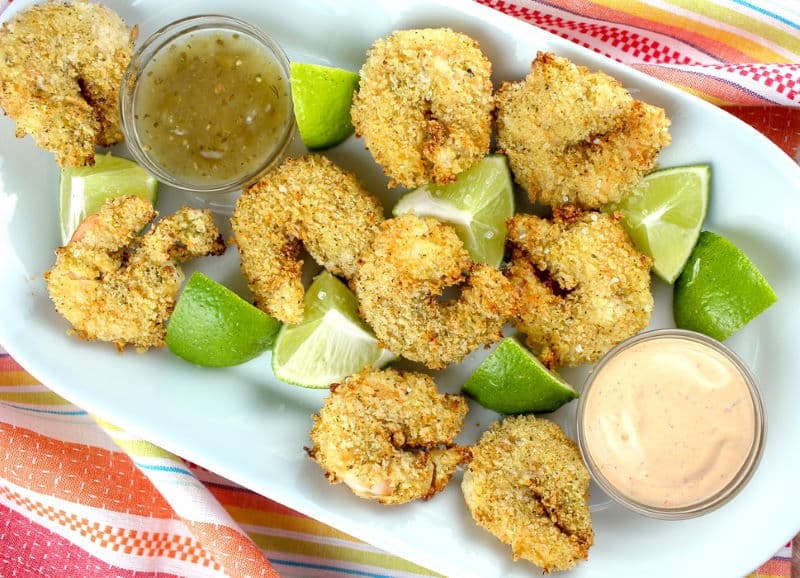 Breaded shrimp with lime wedges and dips in white dish.