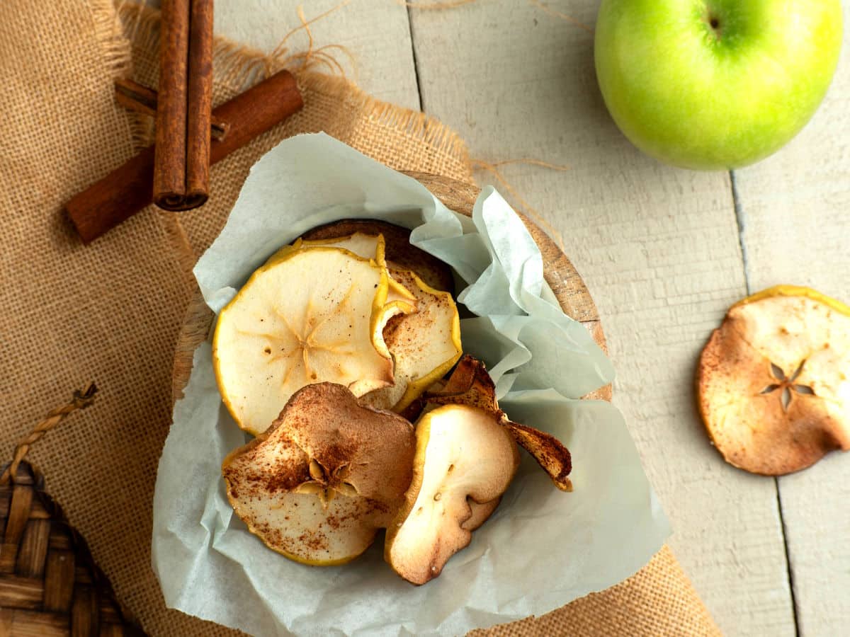 Cinnamon Apple Chips in a wooden bowl with a green apple and cinnamon sticks on the side.