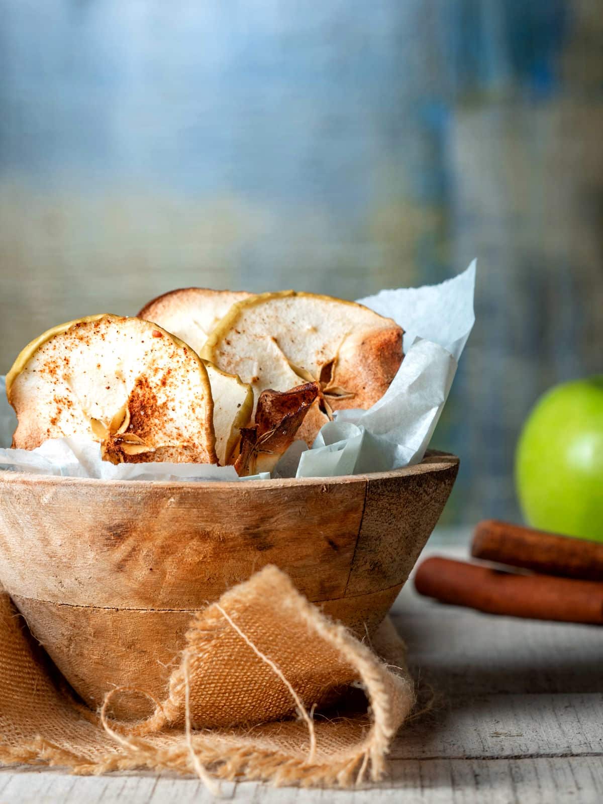 Cinnamon Apple Chips in a wooden bowl with a green apple and cinnamon sticks in the background.