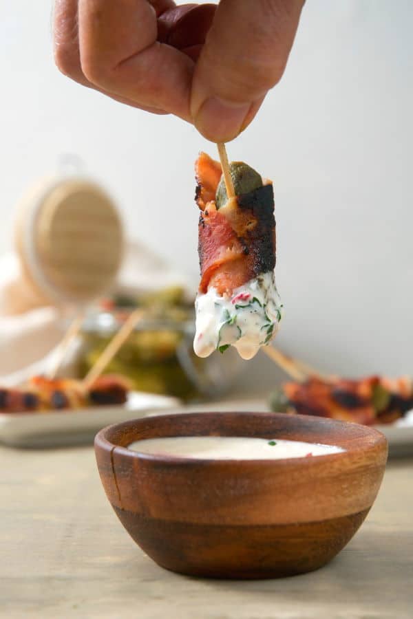 Grilled, bacon wrapped pickle on a toothpick with white dipping sauce in a wooden bowl.