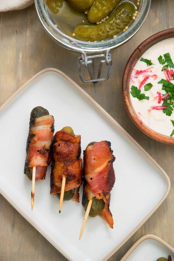 Grilled, bacon wrapped pickles on toothpicks, on a white plate, dipping sauce in the background.