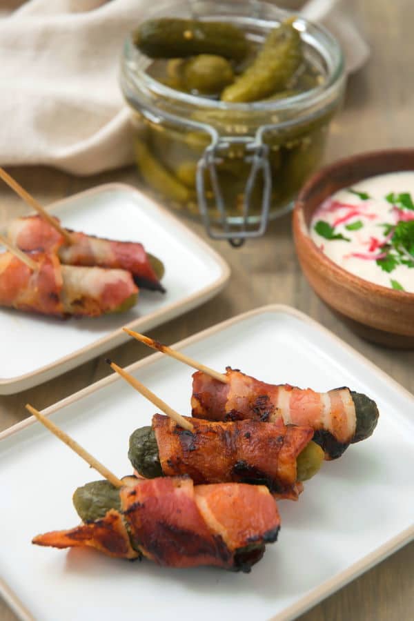 Grilled, bacon wrapped pickles on toothpicks, on a white plate, dipping sauce in the background.