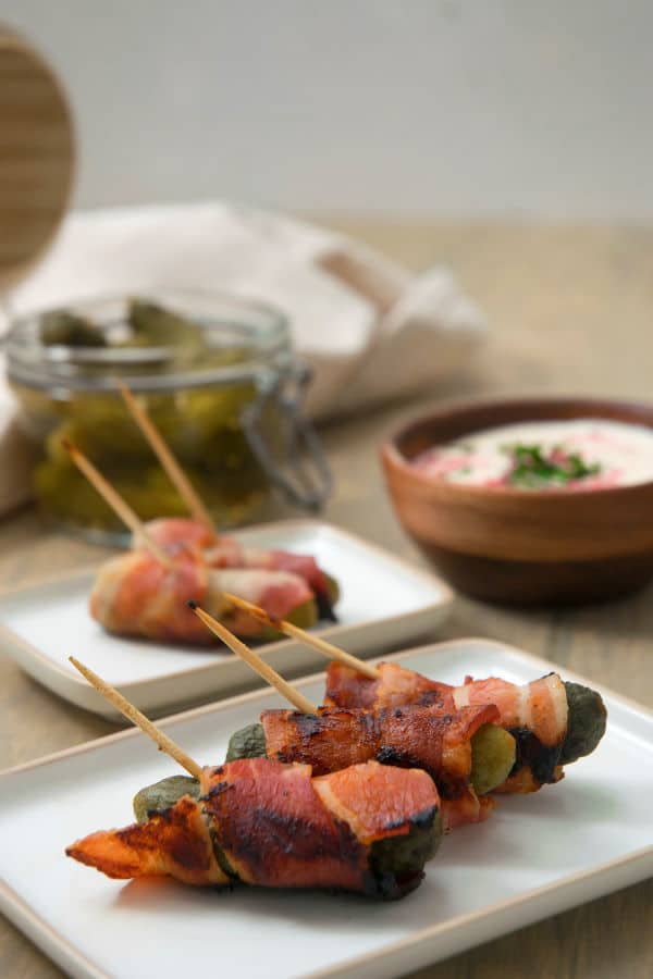 Grilled, bacon wrapped pickles on toothpicks, on a white plate, dipping sauce in the background.