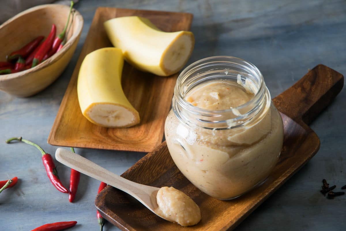 Banana ketchup in a small jar and on a small spoon. Chili peppers and banana pieces on the side.