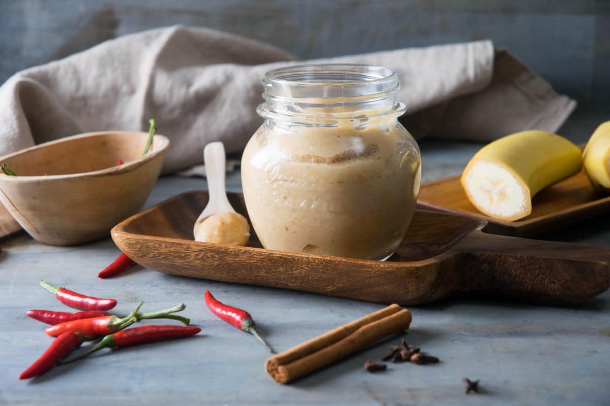 Banana ketchup in a small jar and on a small spoon. Chili peppers, a cinnamon stick and banana pieces on the side.