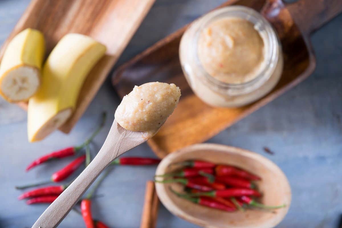 Close up of banana ketchup on a small wooden spoon. Bananas, chili peppers and banana ketchup in a jar in the background unfocused.