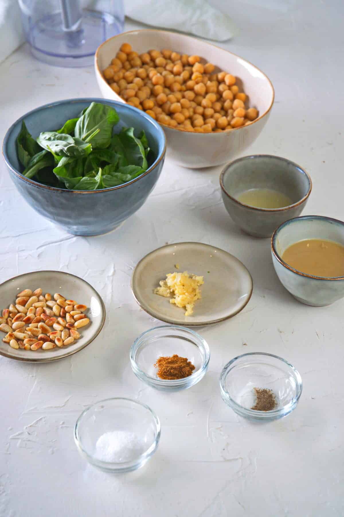 Basil hummus ingredients prepped in bowls on white concrete-look background.