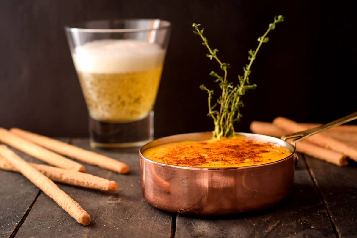 Beer cheese dip in a copper pot with bread sticks and a glass of beer on the side.