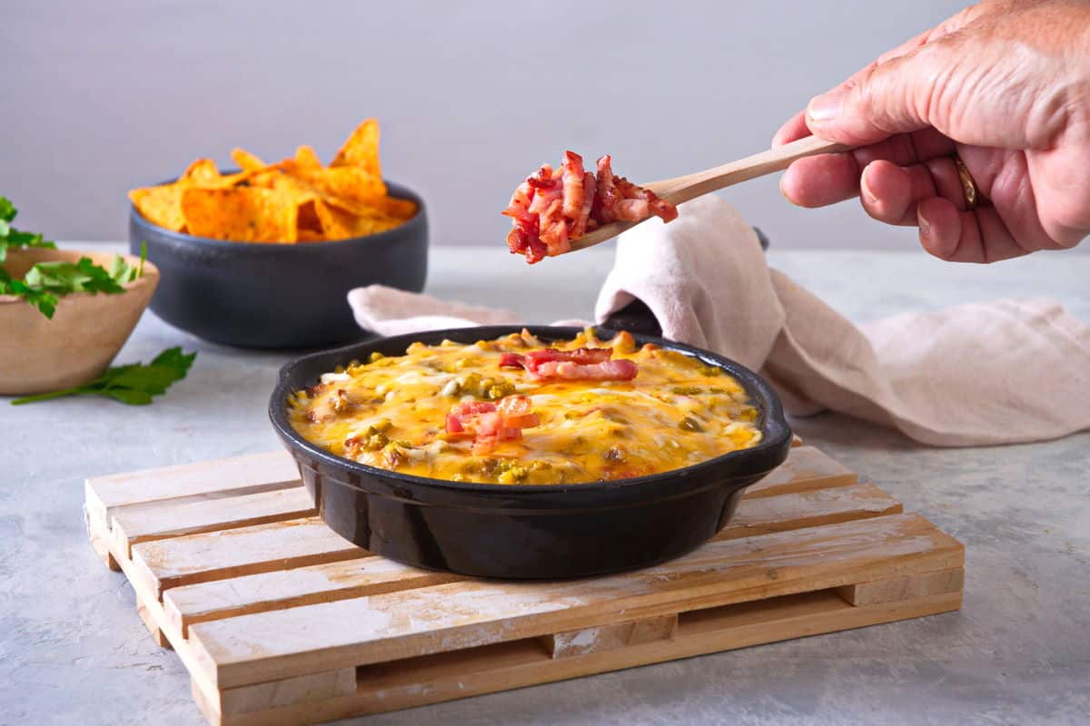 Bison dip in cast iron skillet with bacon bits and a bowl of chips in the background.