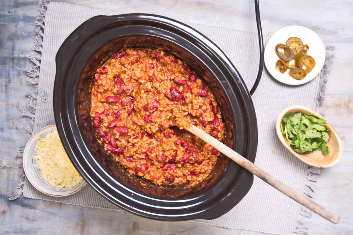 Bison chili in slow cooker with cilantro, cheese and jalapeno slices on the side.