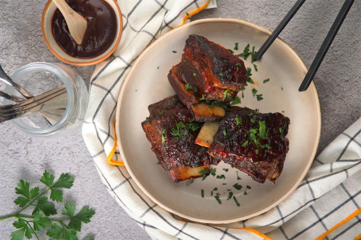 Cooked BBQ bison ribs in white dish with a kitchen towel on the side.