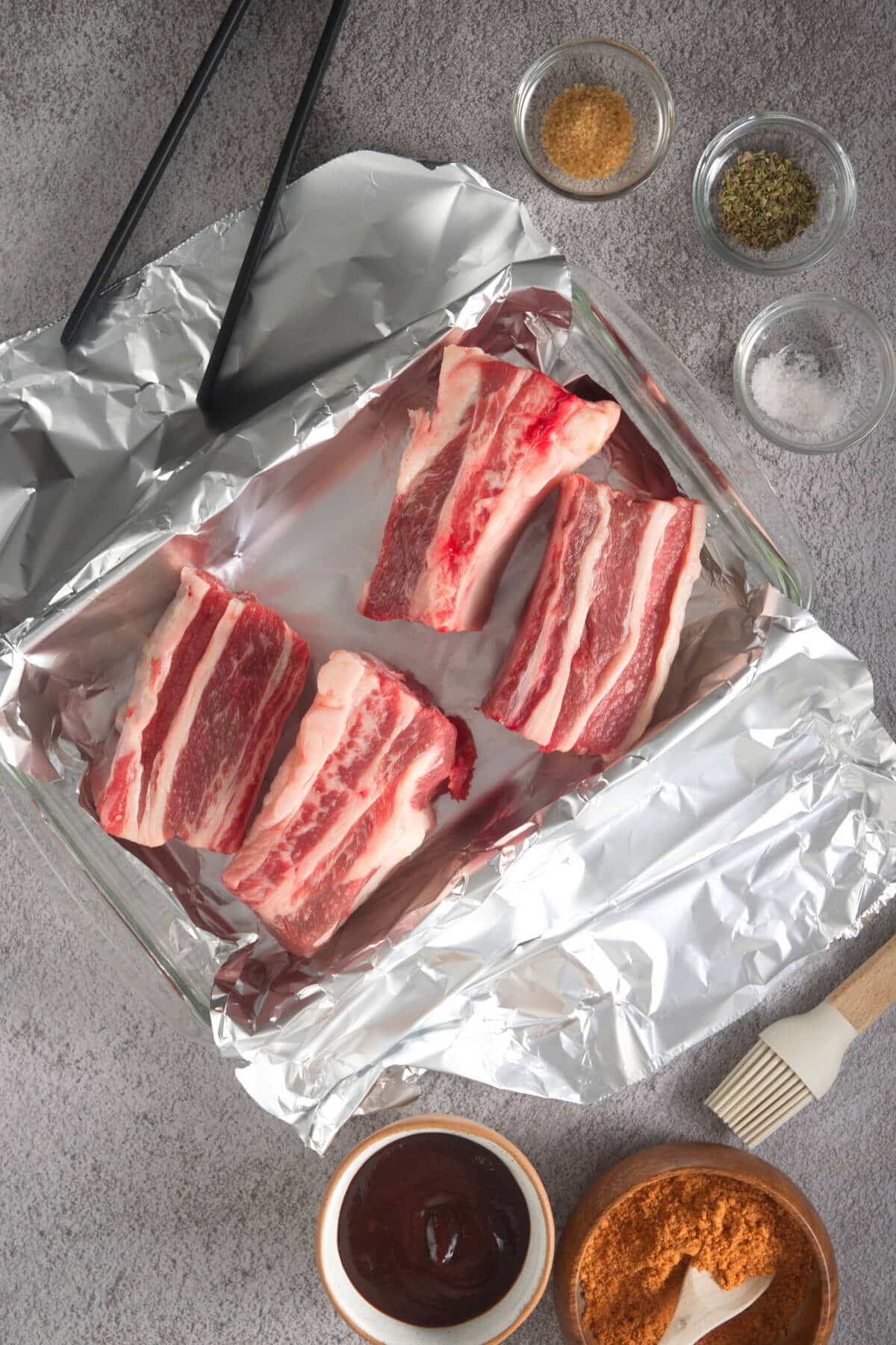 Raw bison short ribs in foil lined baking dish with spices on the side.