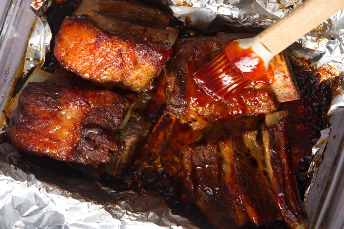 Bison short ribs getting covered with BBQ sauce on foil.