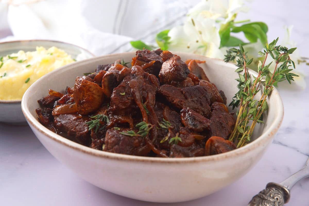 Bison stew with fresh thyme in a light-colored bowl, mashed potatoes and white flowers in the background.