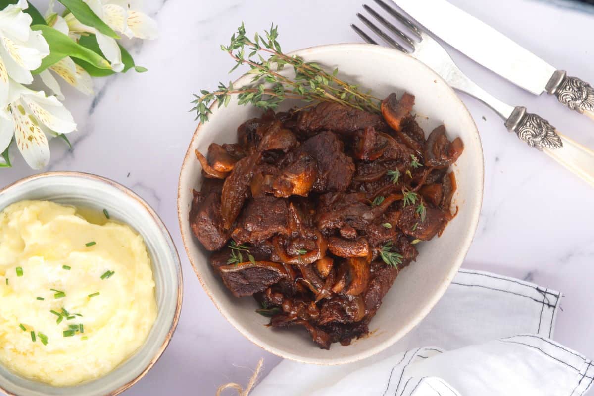 Bison stew with fresh thyme in a light-colored bowl, mashed potatoes and white flowers on the side.