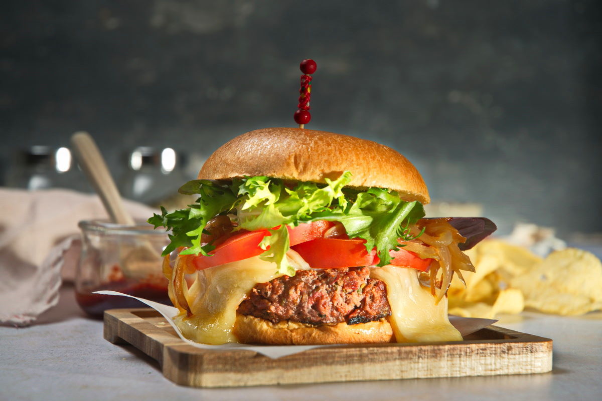 Bison burger with lettuce, tomato and melted cheese on wooden cutting board.