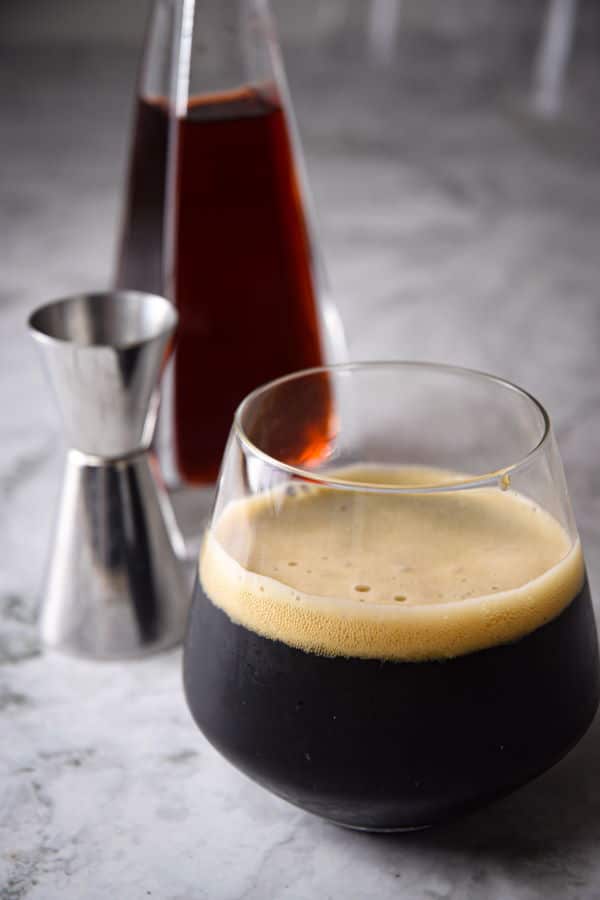 Black fog cocktail in a glass, raspberry liqueur in the background, on marble.