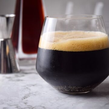 Black fog cocktail in a glass, raspberry liqueur in the background, on marble.