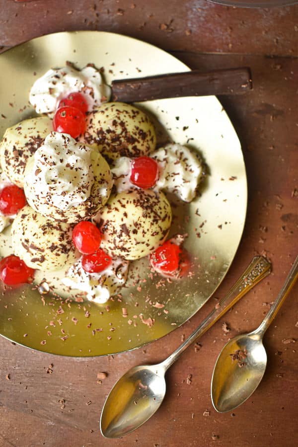 Black forest cake balls on a plate with whipped topping, cherries and chocolate shavings, spoons on the side.