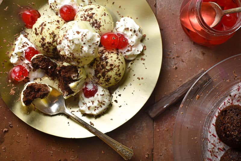 Black forest cake balls on a plate with whipped topping, cherries and chocolate shavings, cherries on the side.