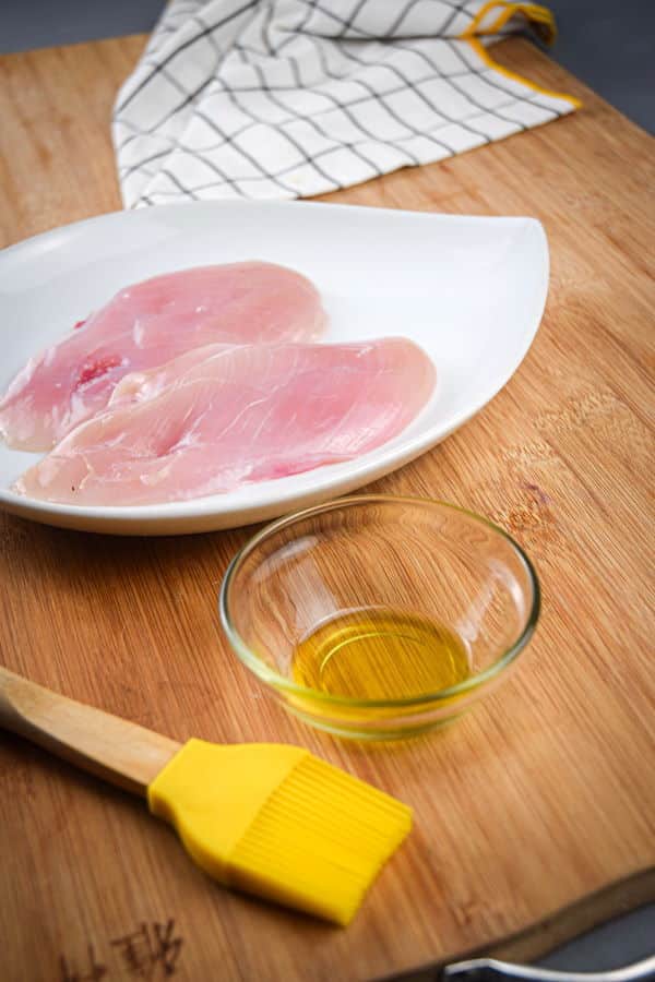 Raw chicken breast on a plate and oil in a small bowl, silicone brush on the side.