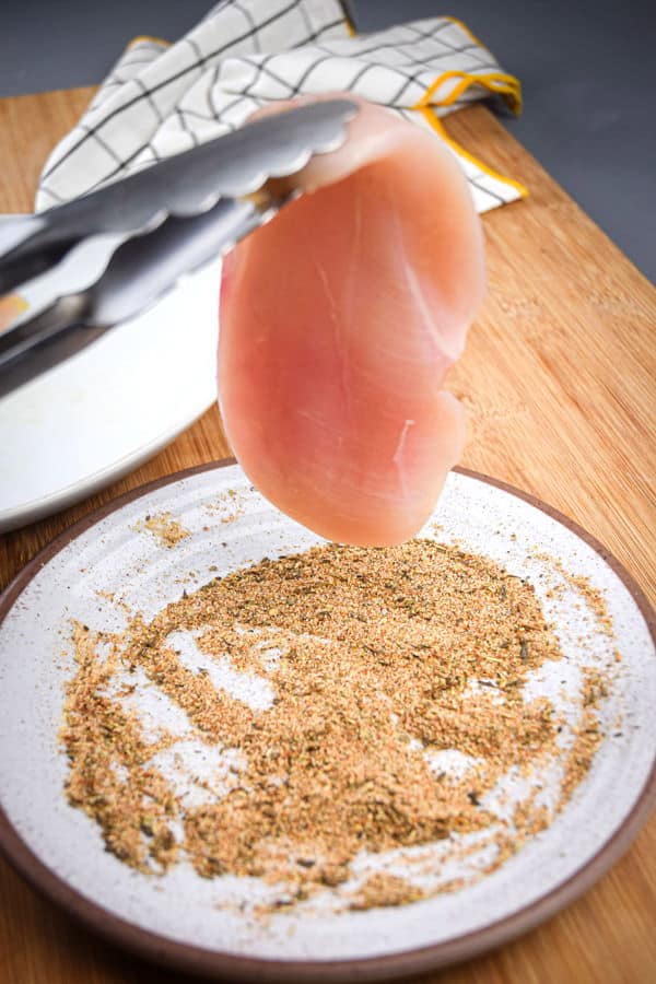 Tongs holding a raw chicken with spices on a plate.